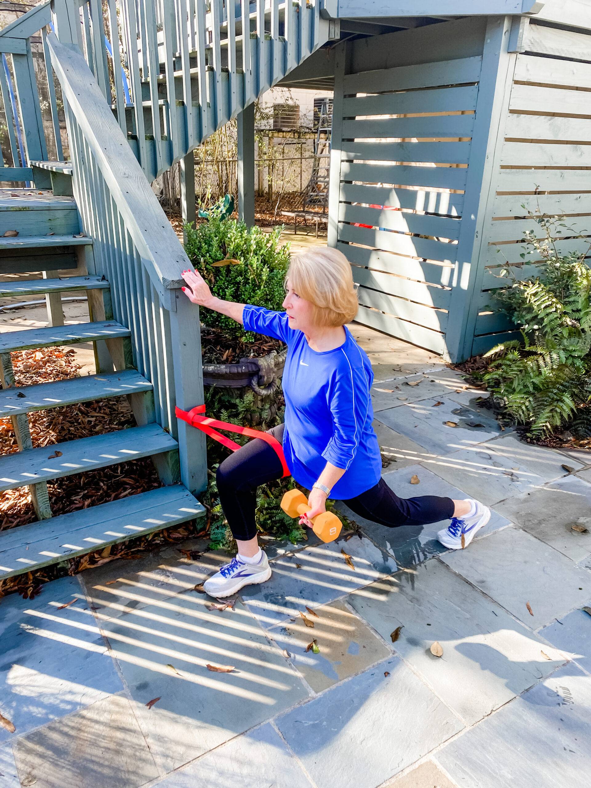 A women performs a lunge exercise outdoors using a resistance band anchored to the staircase for support. She holds a dumbbell in one hand and is stabilizing herself with the other hand on the railing.