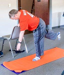 An older adult with intellectual and physical disabilities practices a modified Warrior III pose using a chair for support in an adaptive yoga class. Adaptwell Fitness promotes accessible yoga and fitness for all abilities.